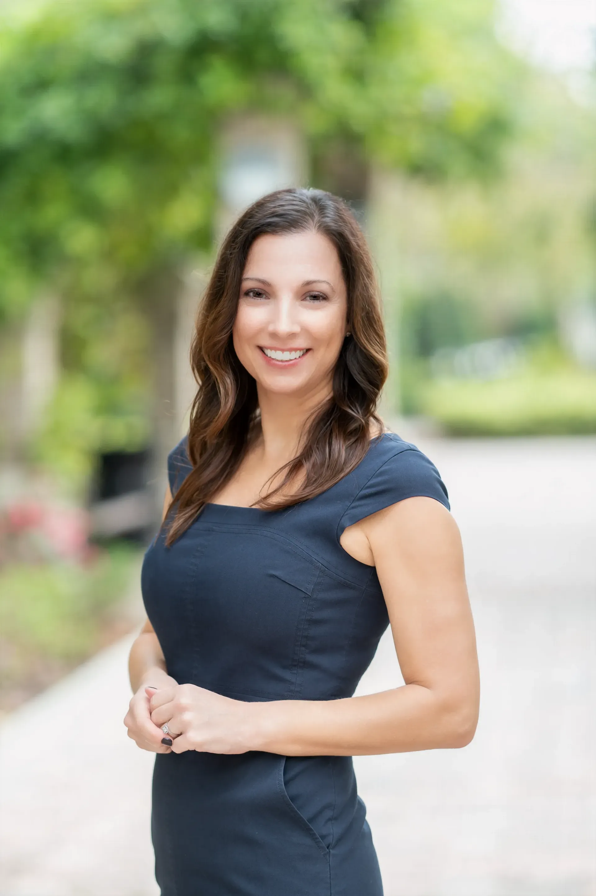 Woman in a navy dress smiles, standing outdoors near a walkway with greenery.