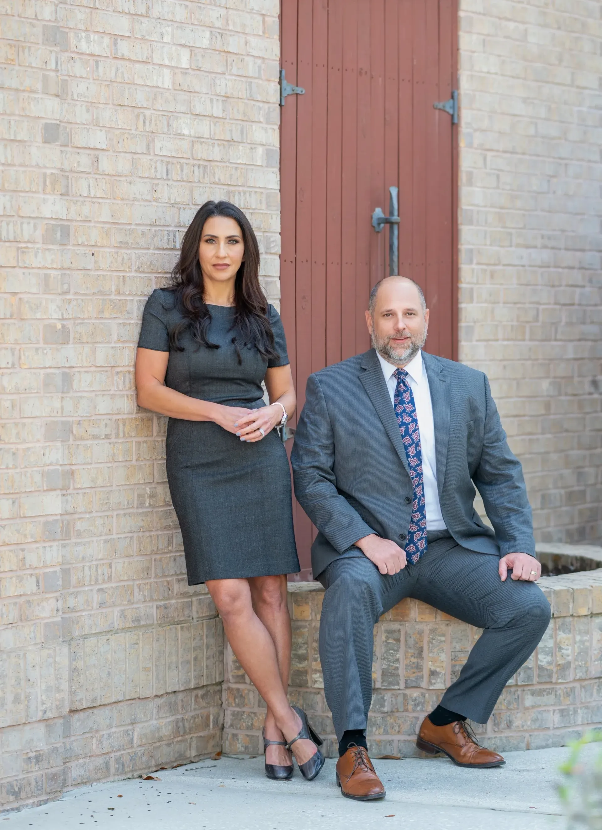 Woman in gray dress leans against brick wall; man in suit sits nearby.
