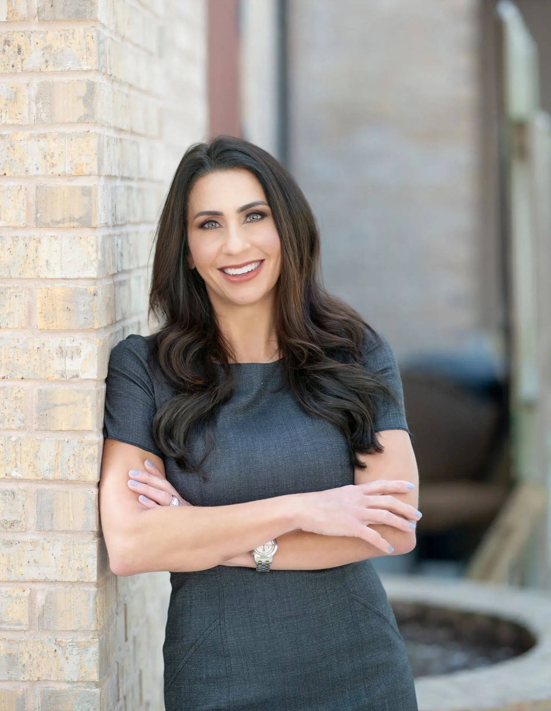 Woman with long dark hair, wearing a sparkly gray dress, smiling, leaning against a brick wall, arms crossed.