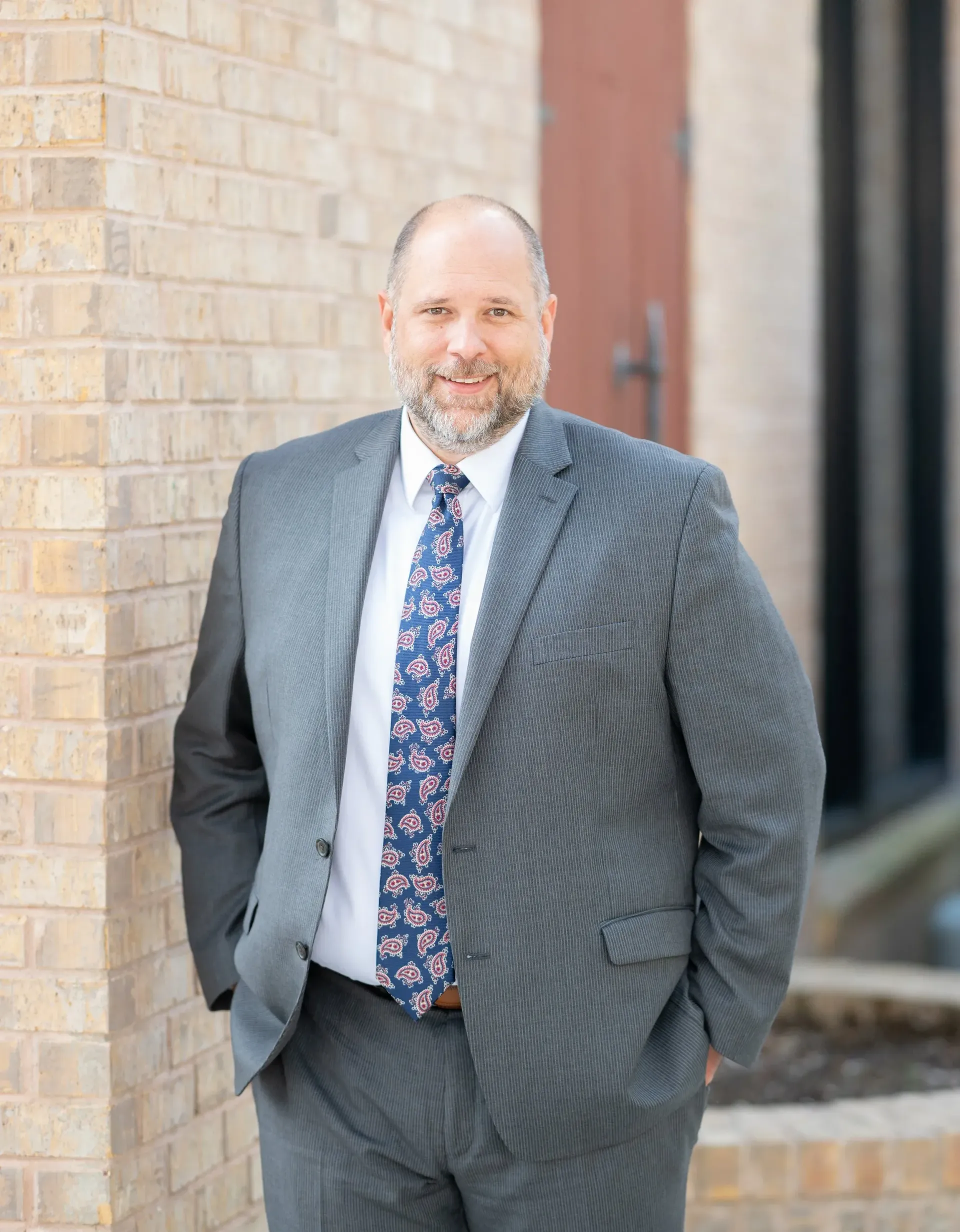 Man in suit leaning against a brick wall, looking at the camera with a slight smile.