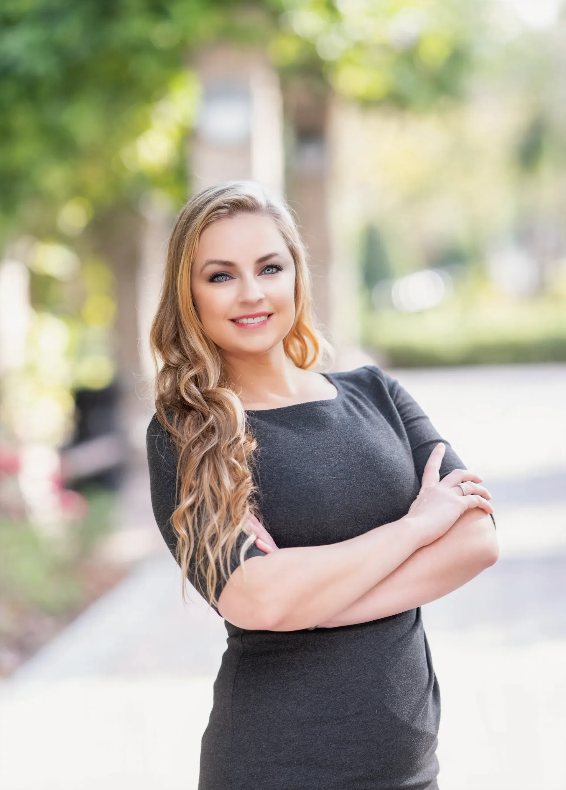 Woman with blonde hair, arms crossed, wearing a gray dress, smiling outdoors.