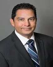 Man in suit and tie smiling, posing for a portrait against a gray backdrop.