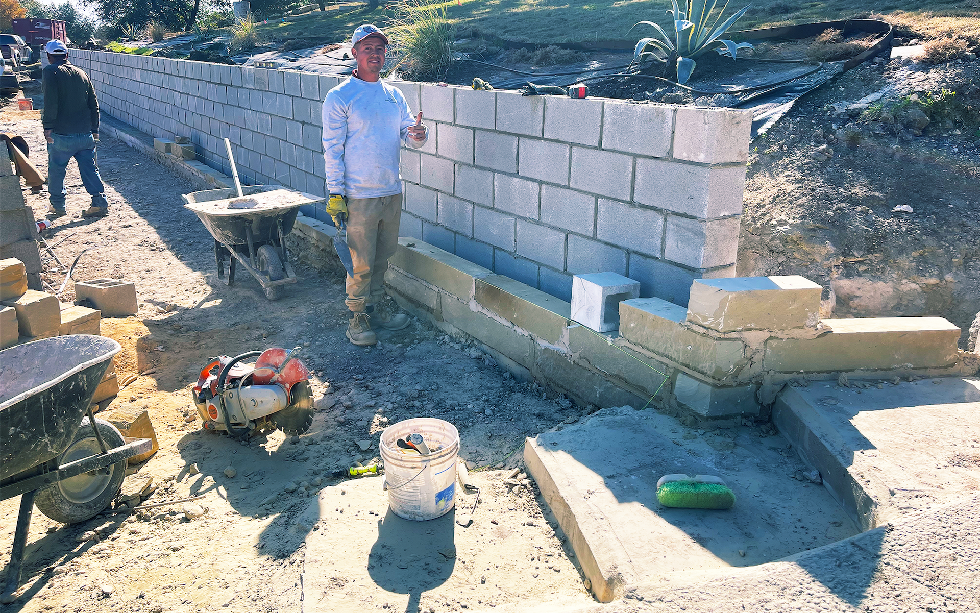 Man building a cinder block wall outdoors, giving a thumbs up. Other workers nearby.