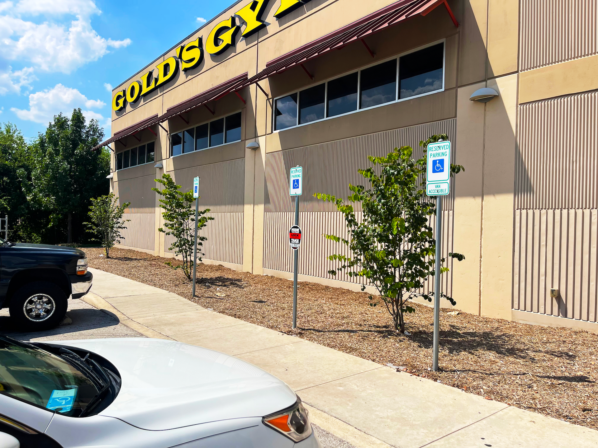Gold's Gym building with accessible parking signs and landscaping. Sunny day, cars parked out front.