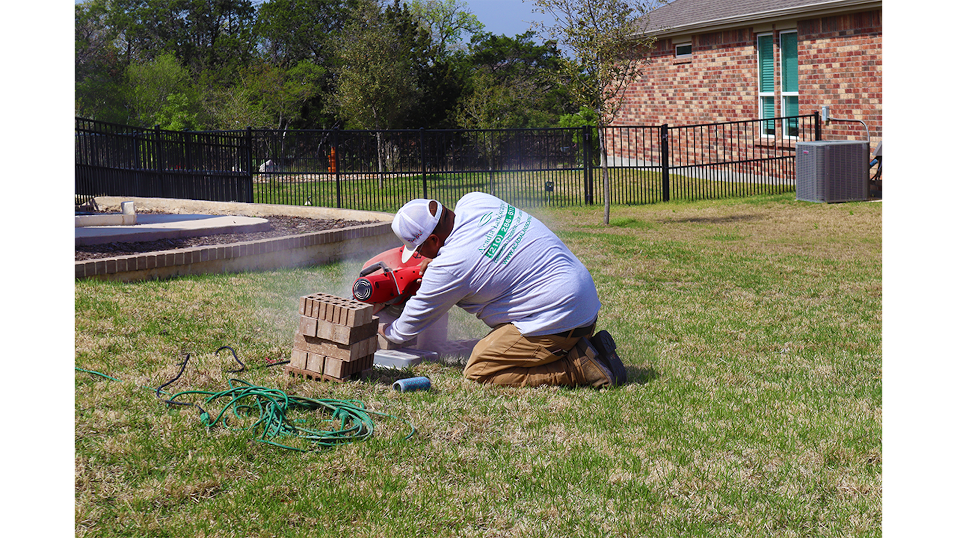 Man in white shirt kneeling, cutting bricks with a saw on a lawn. Brick home and fence in background.