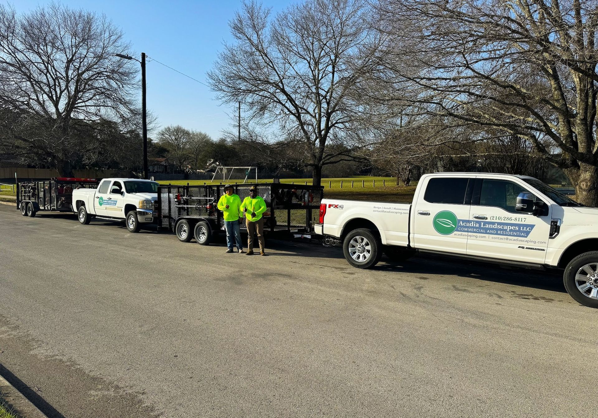 Two white trucks with trailers and two people wearing bright vests stand on a road near trees.