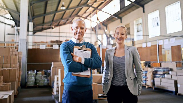 Two warehouse workers smiling in a storage facility, one holding a clipboard with boxes and pallets behind them