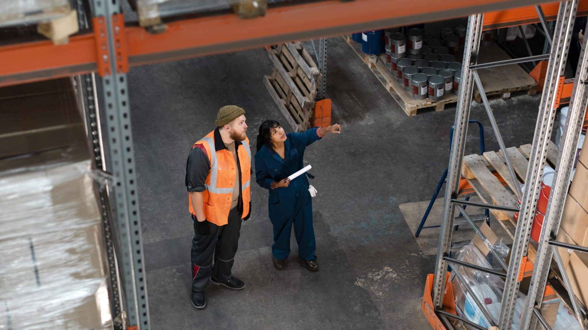Woman in coveralls pointing out area of interest in warehouse to man wearing safety vest