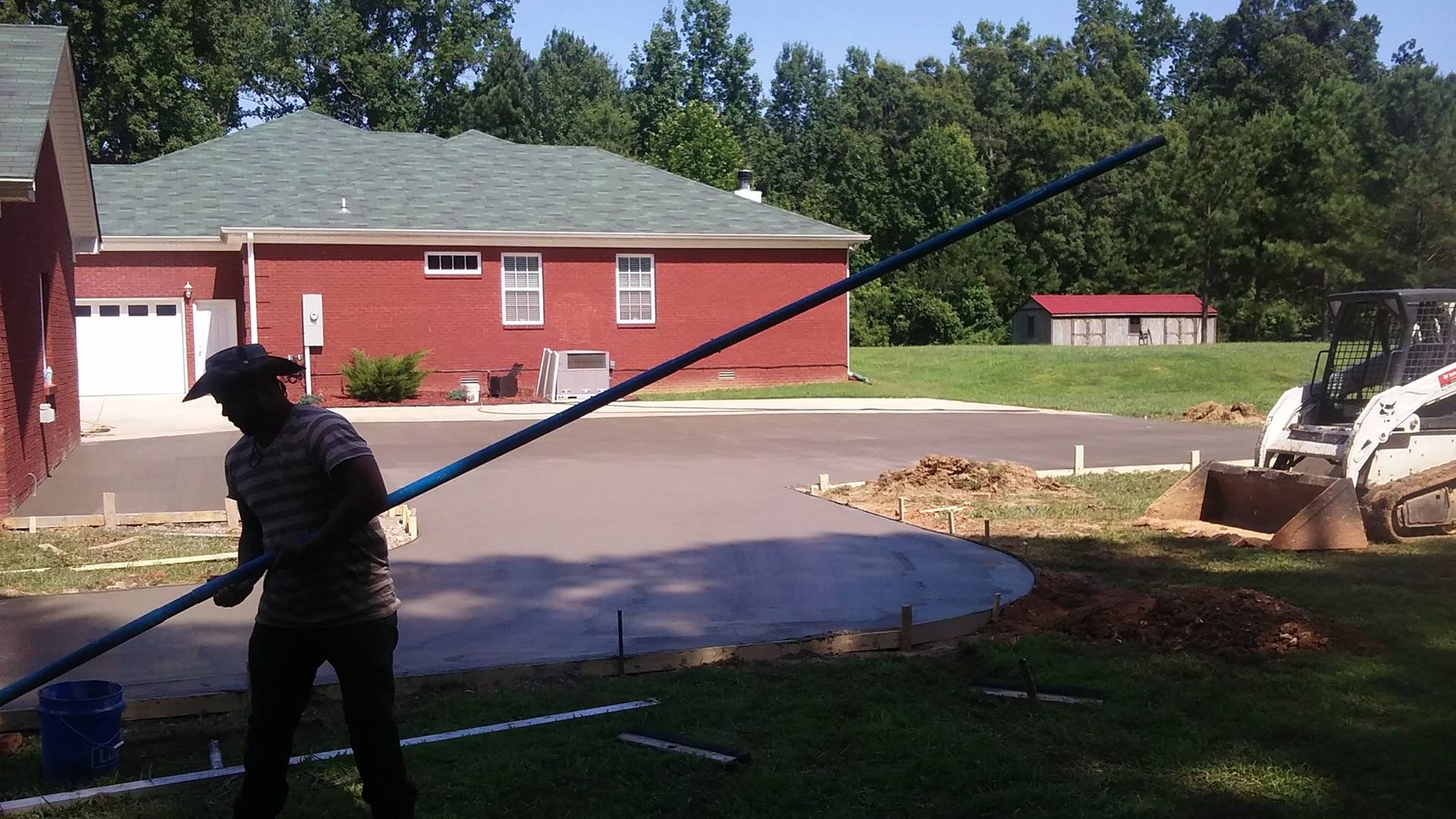 Man smoothing wet concrete driveway with a long tool; house and trees in background.