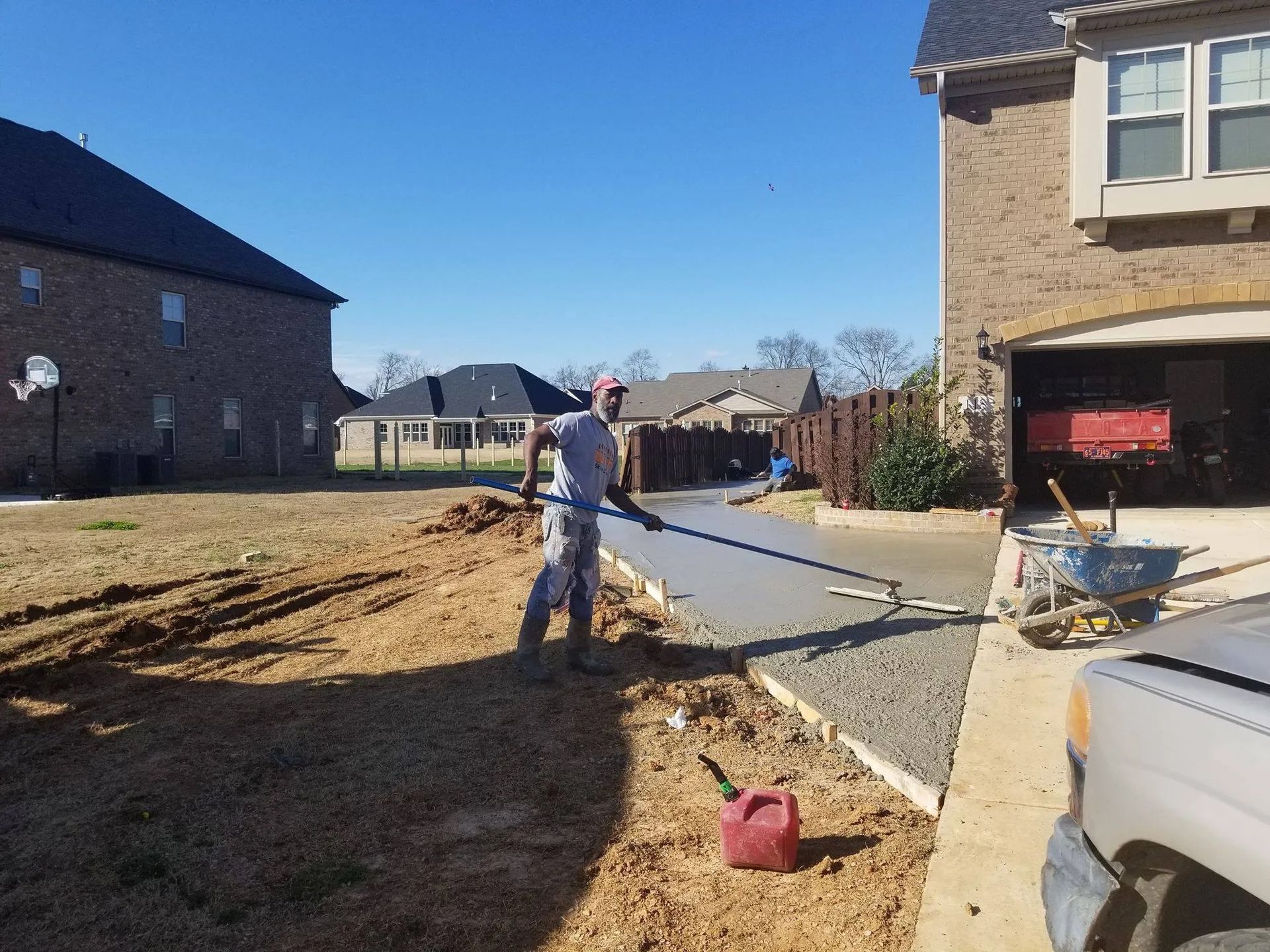 Person leveling wet concrete with a long tool on a driveway, near a house and lawn on a sunny day.