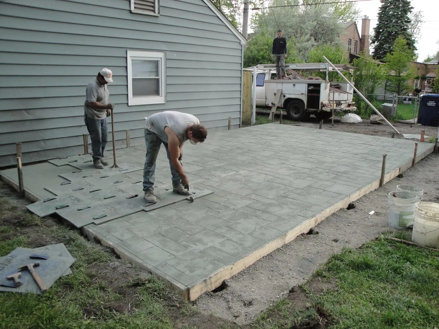 Workers leveling concrete for a patio next to a light blue house, with a truck in the background.