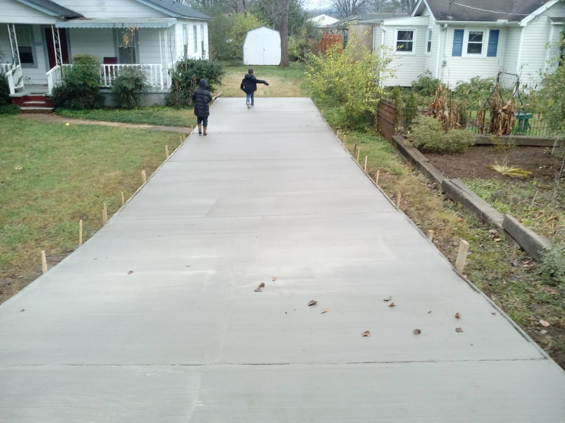 Newly poured concrete driveway with two people walking on it, flanked by houses and wooden stakes.