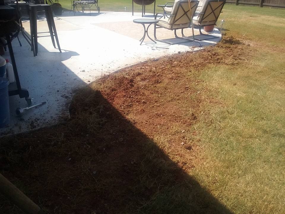 A patch of soil bordering a concrete patio and lawn. Dirt and grass are side by side in a sunny outdoor setting.