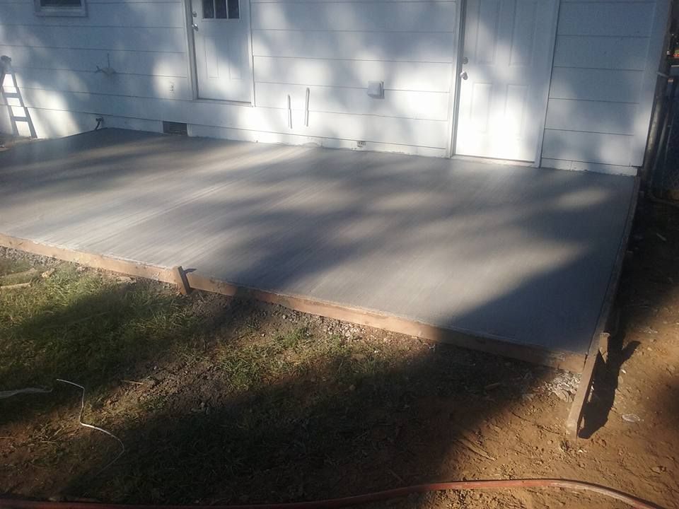 A newly poured concrete patio outside a white building. The patio is surrounded by grass and dirt.