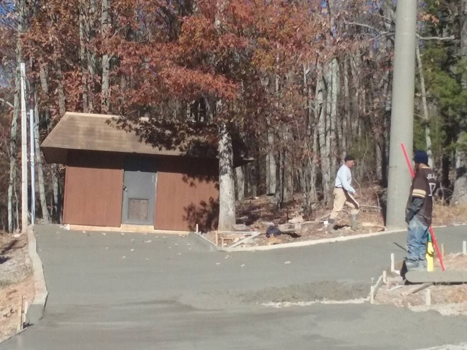 Two men working on a concrete driveway near a small brown building and trees.