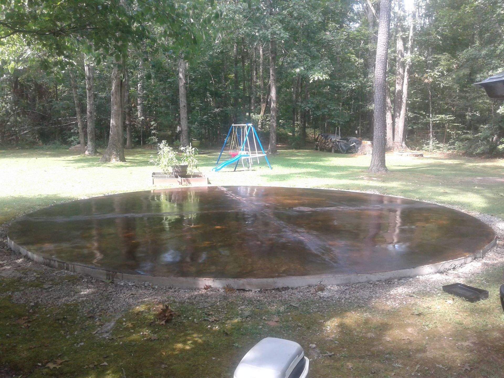 Circular concrete area filled with water, surrounded by gravel, in a grassy backyard with trees and a playset in the background.