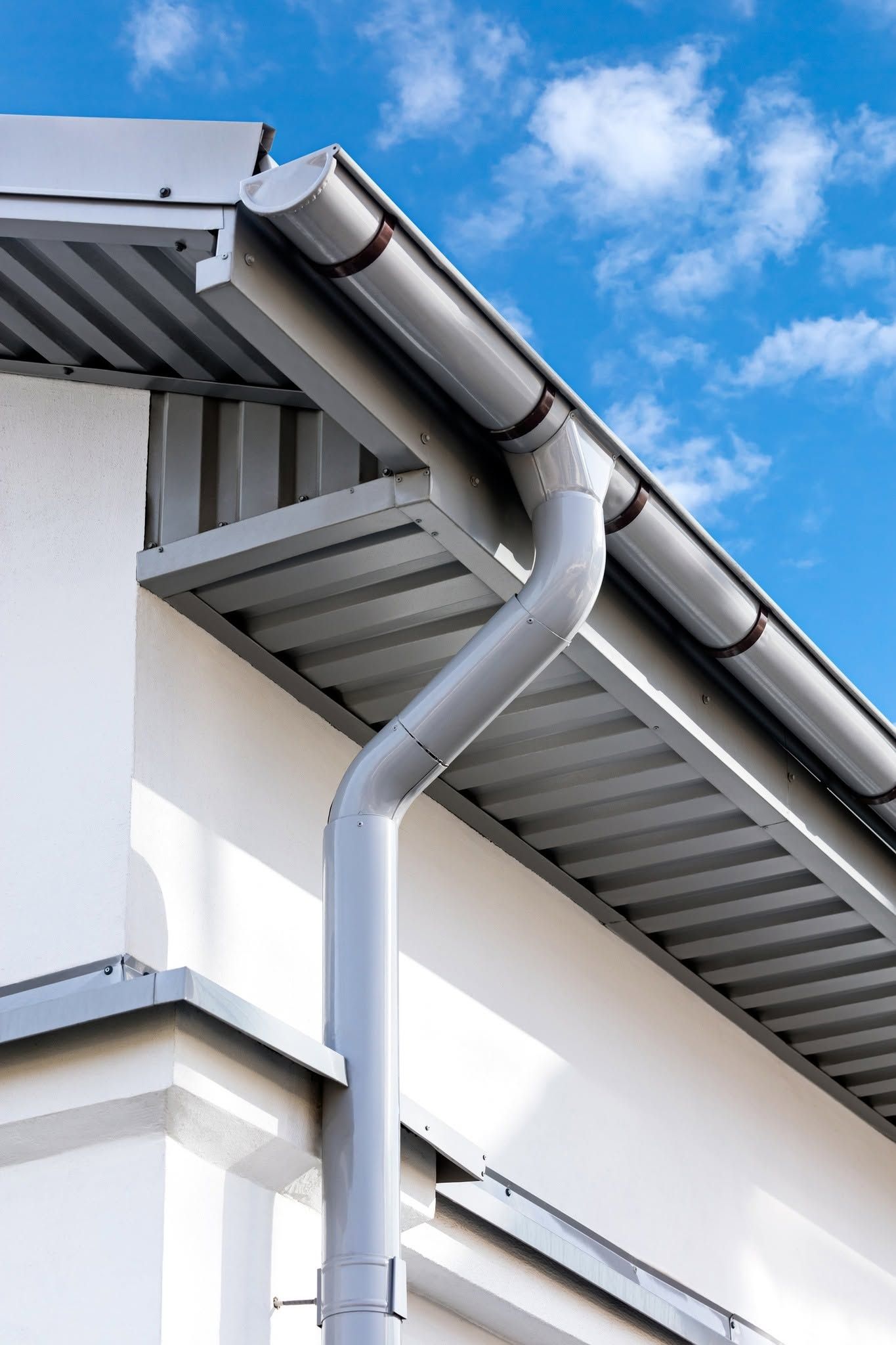 White downspout on a house under a gray gutter against a blue sky with clouds
