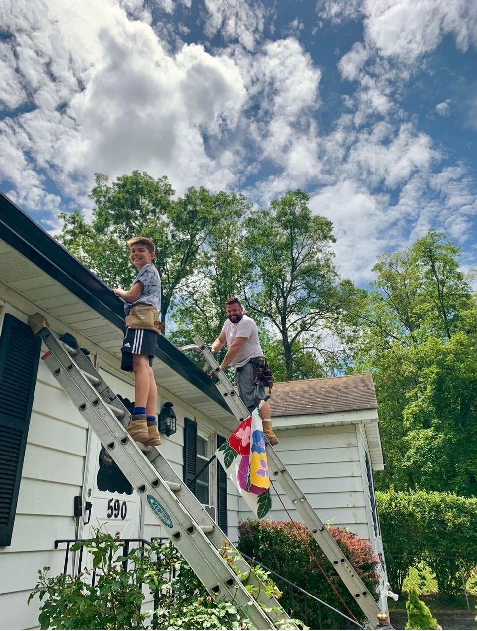 Two workers on a roof repairing siding and gutters beside a white house under a blue sky