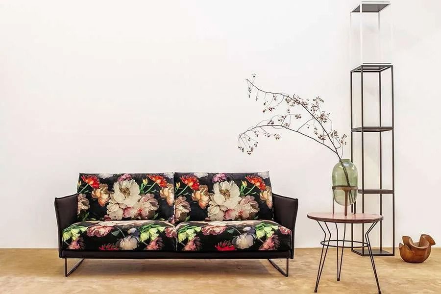 Divano con stampa floreale in una stanza bianca con tavolino laterale in metallo, mensola e un vaso con rami.