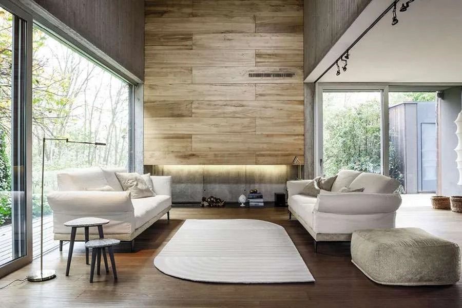 Soggiorno moderno con divani dai colori chiari, una parete decorativa in legno, ampie finestre e un tappeto.