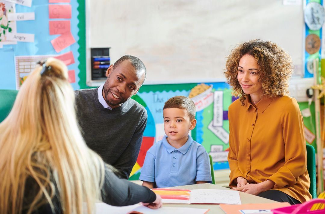 A teacher, parents, and a child are seated around a table in a classroom, discussing something.