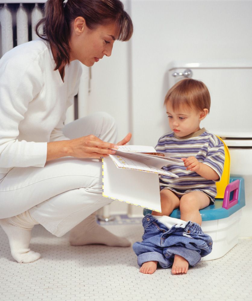 Woman reading a book to a toddler seated on a potty chair in a playroom.
