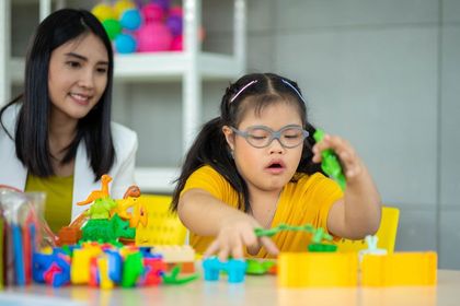 Woman assisting child with Down syndrome playing with colorful toys at a table.
