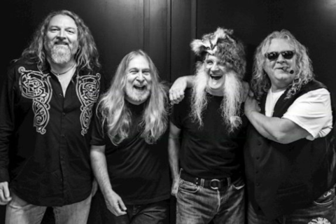 Four long-haired men posing in a black and white photo. One wears a hat with a beard, another smokes a cigar.