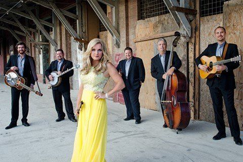 Bluegrass band poses: woman in yellow dress, men in suits with instruments in an urban setting.