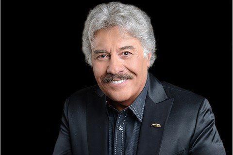 Smiling, older man with gray hair and mustache wearing a black suit jacket against a black background.