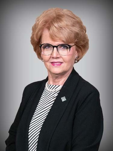A woman wearing glasses and a black jacket is standing in front of a gray background.