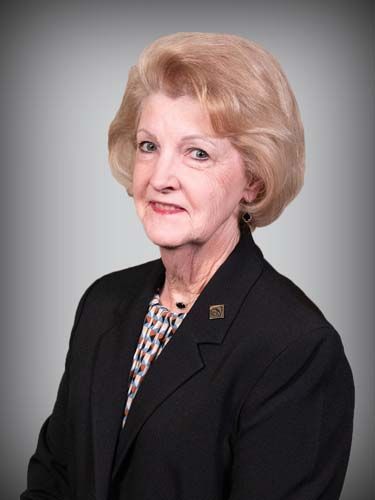 A woman wearing a black jacket and a brooch is standing in front of a gray background.