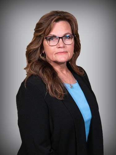 A woman wearing glasses and a black jacket is standing in front of a gray background.