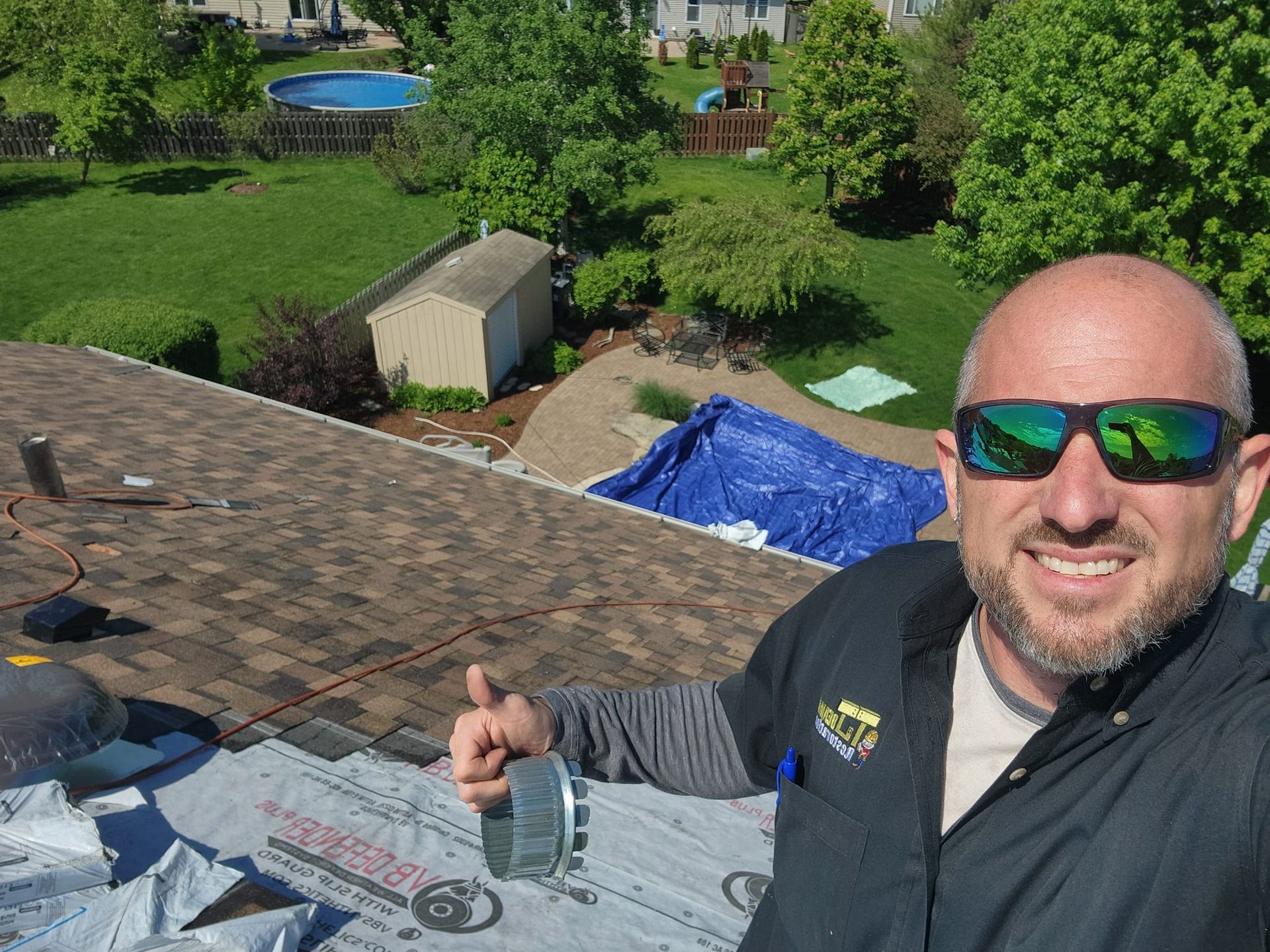 A man wearing sunglasses is standing on top of a roof giving a thumbs up.