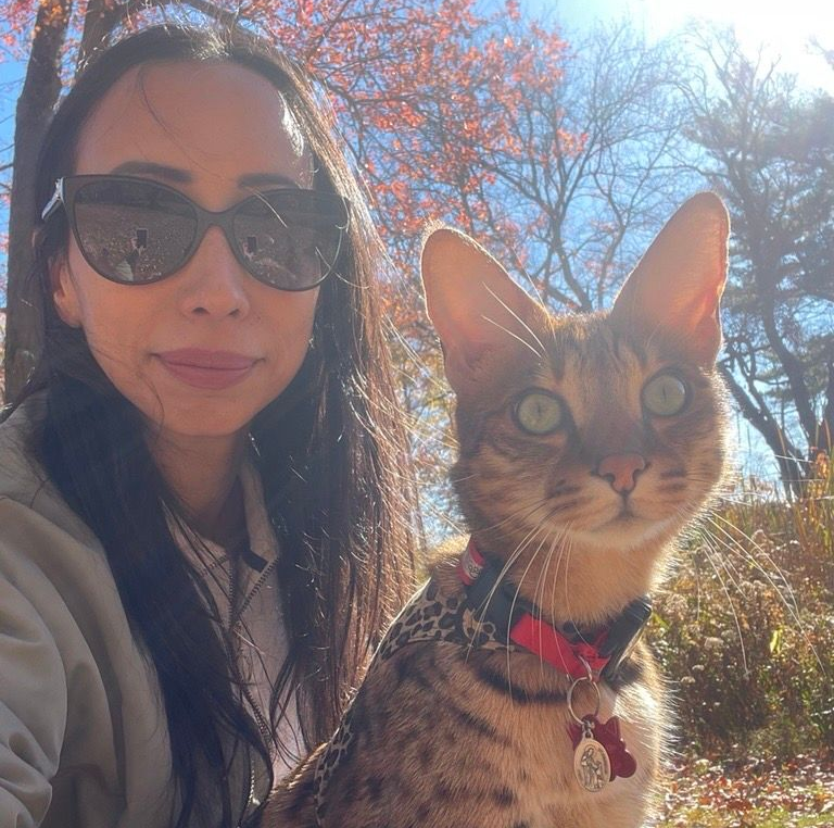A person with dark hair and sunglasses poses outdoors with a spotted cat wearing a patterned harness and a red collar.