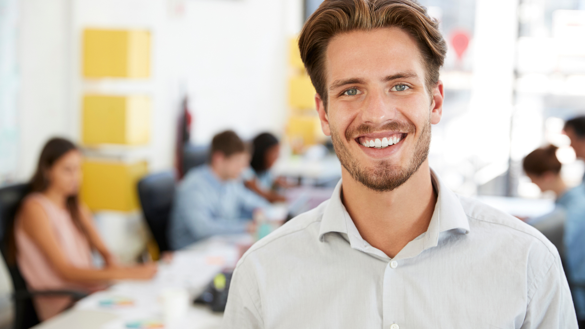 Man smiling at camera in modern office with colleagues in the background.