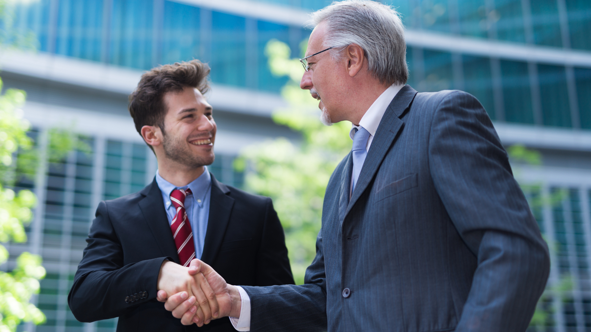 Two men in suits shaking hands outside a building; one is smiling.