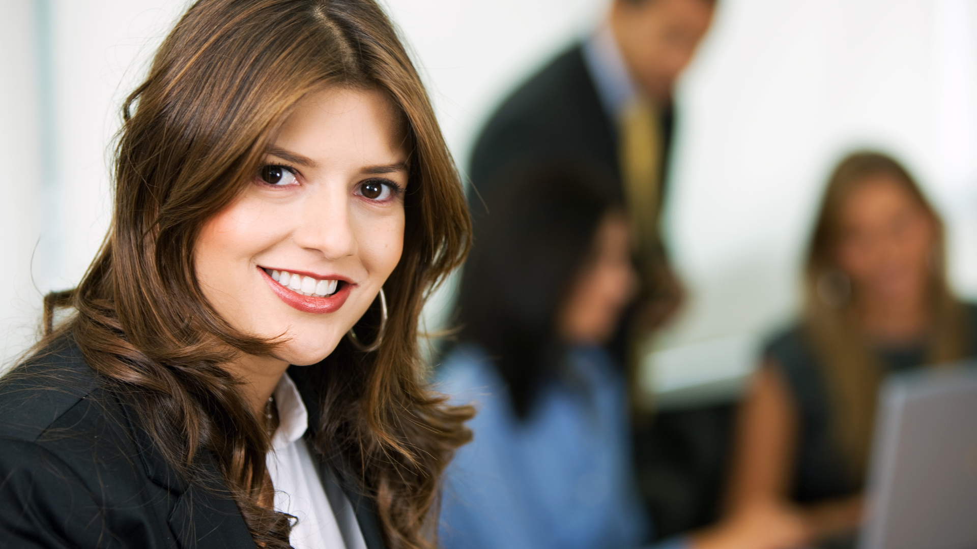 Woman in a blazer smiles at the camera, others in the office setting blurred in the background.