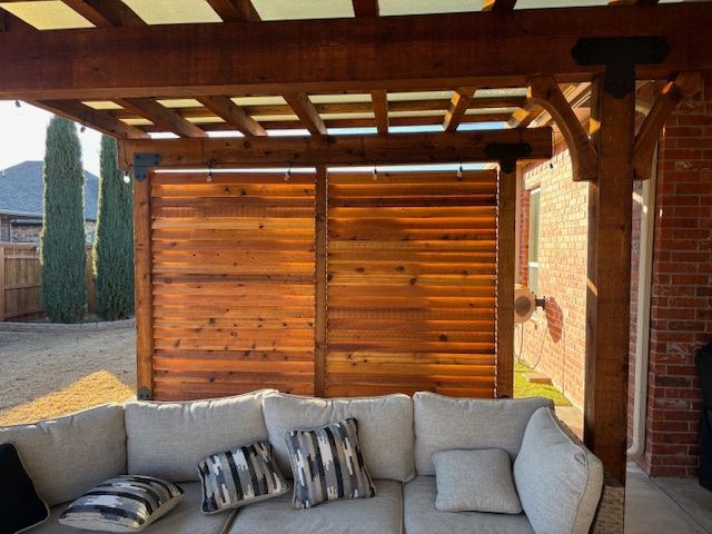 A couch under a wooden pergola with a brick building in the background