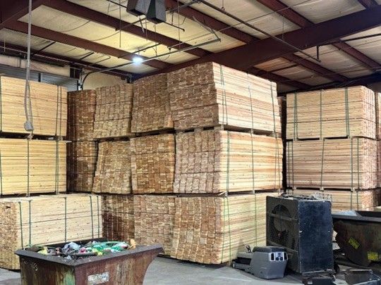 Stacks of bundled lumber neatly piled on pallets inside a large warehouse with exposed steel beams and industrial equipment visible.