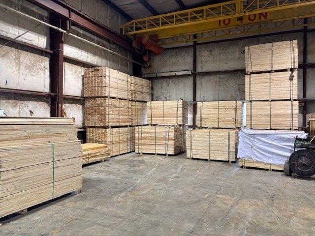 Interior of an industrial warehouse with stacks of bundled lumber neatly arranged on pallets, a yellow overhead crane labeled 10-ton above, and a concrete floor with open space in the foreground