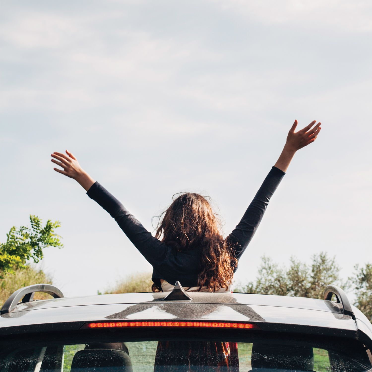 Person with arms raised through sunroof of a car, outdoors under a bright sky near Prelude at Medley.