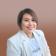 A person with short, dark hair wears a white blazer over a graphic t-shirt, smiling against a solid brown background.