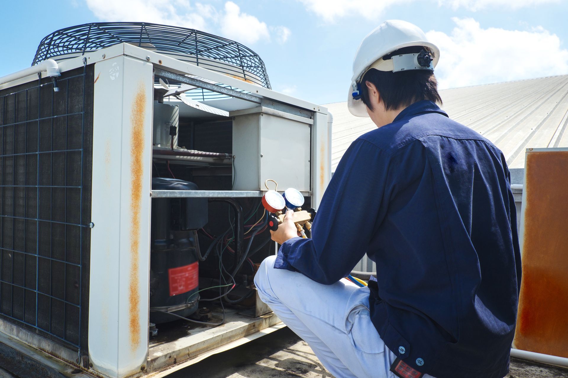 An HVAC technician in a hard hat inspects rooftop air conditioning unit, using gauges. The technician is outdoors on a sunny day.