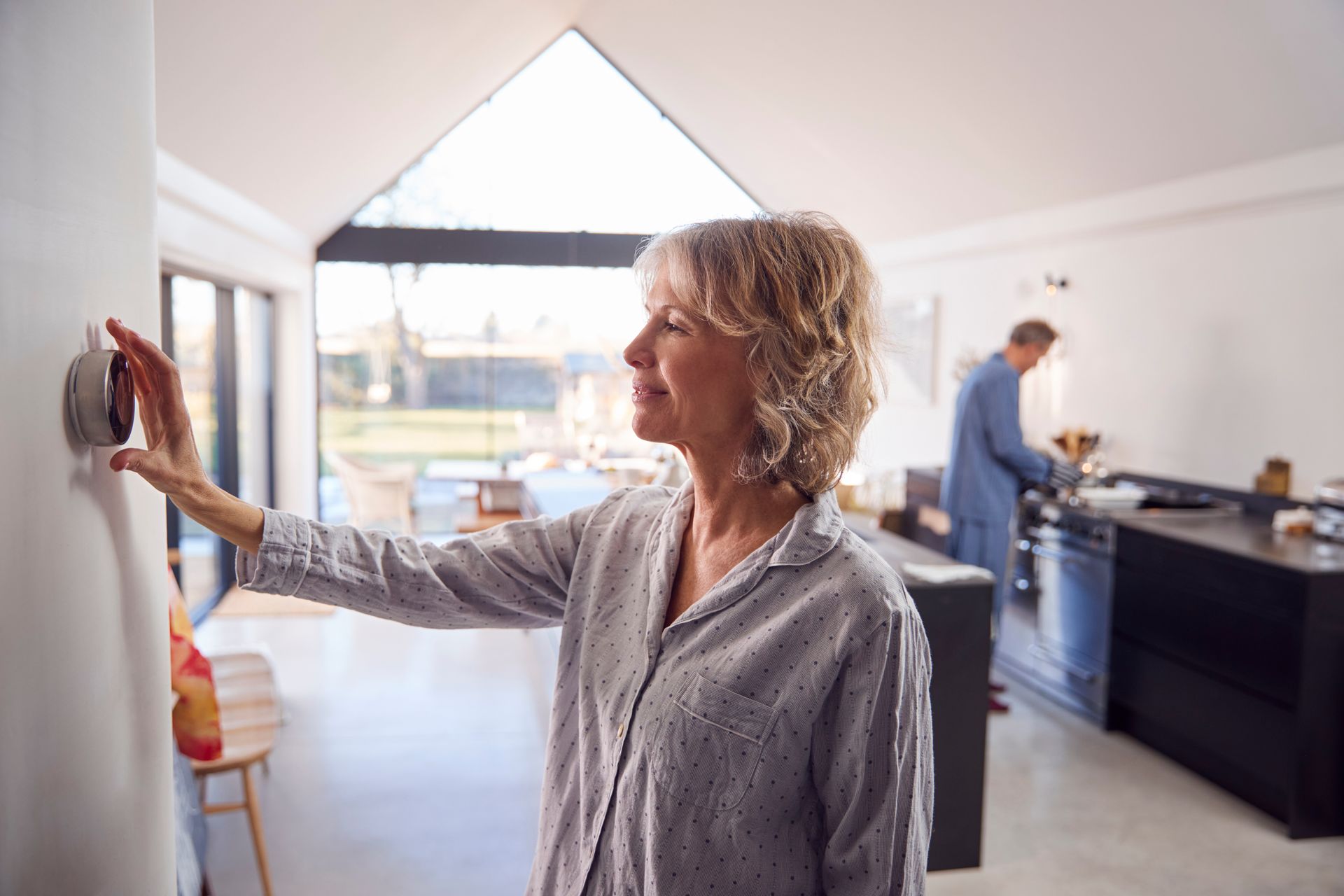 Woman adjusting a thermostat in a modern kitchen, with a man cooking in the background.