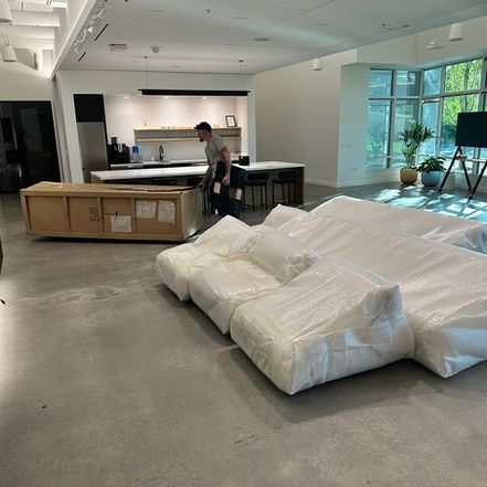 A person in an office moves a wooden crate, near a bubble-wrapped couch. Kitchen and large windows in background.