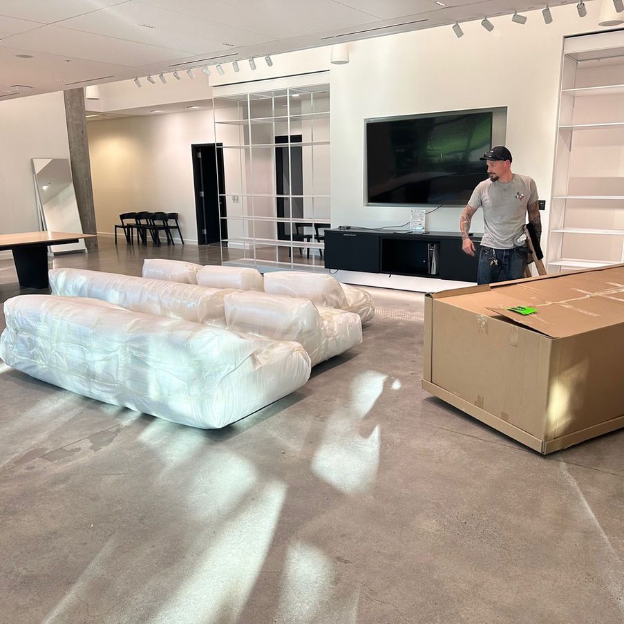 Man unpacks furniture in a mostly empty, modern-style room with a large TV and concrete floor.