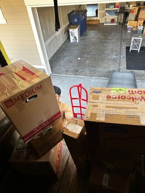 Person in tan shirt with boxes in garage; moving boxes. Red hand truck in background.