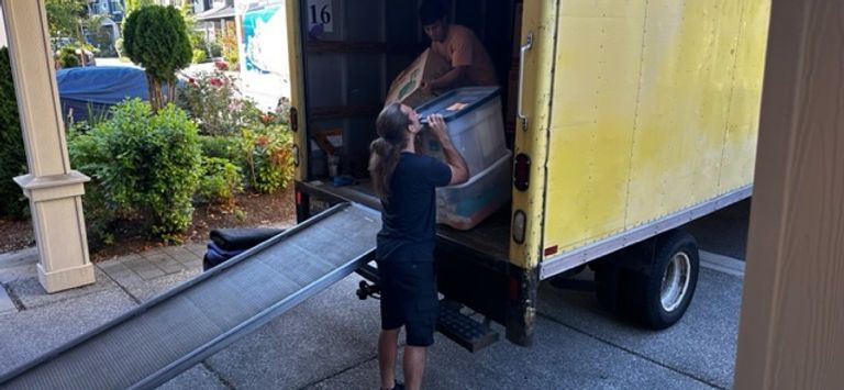 Men loading a large container into the back of a yellow moving truck using a ramp.