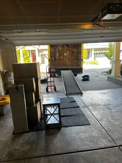 Moving truck ramped down in a garage; boxes stacked, a dolly, and a small table.